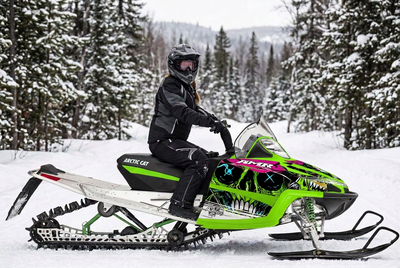 Woman rider seated on an Arctic Cat M Series snowmobile with green, pink and black Frenzy skull graphics and bright green plastics on a snowy forest trail.